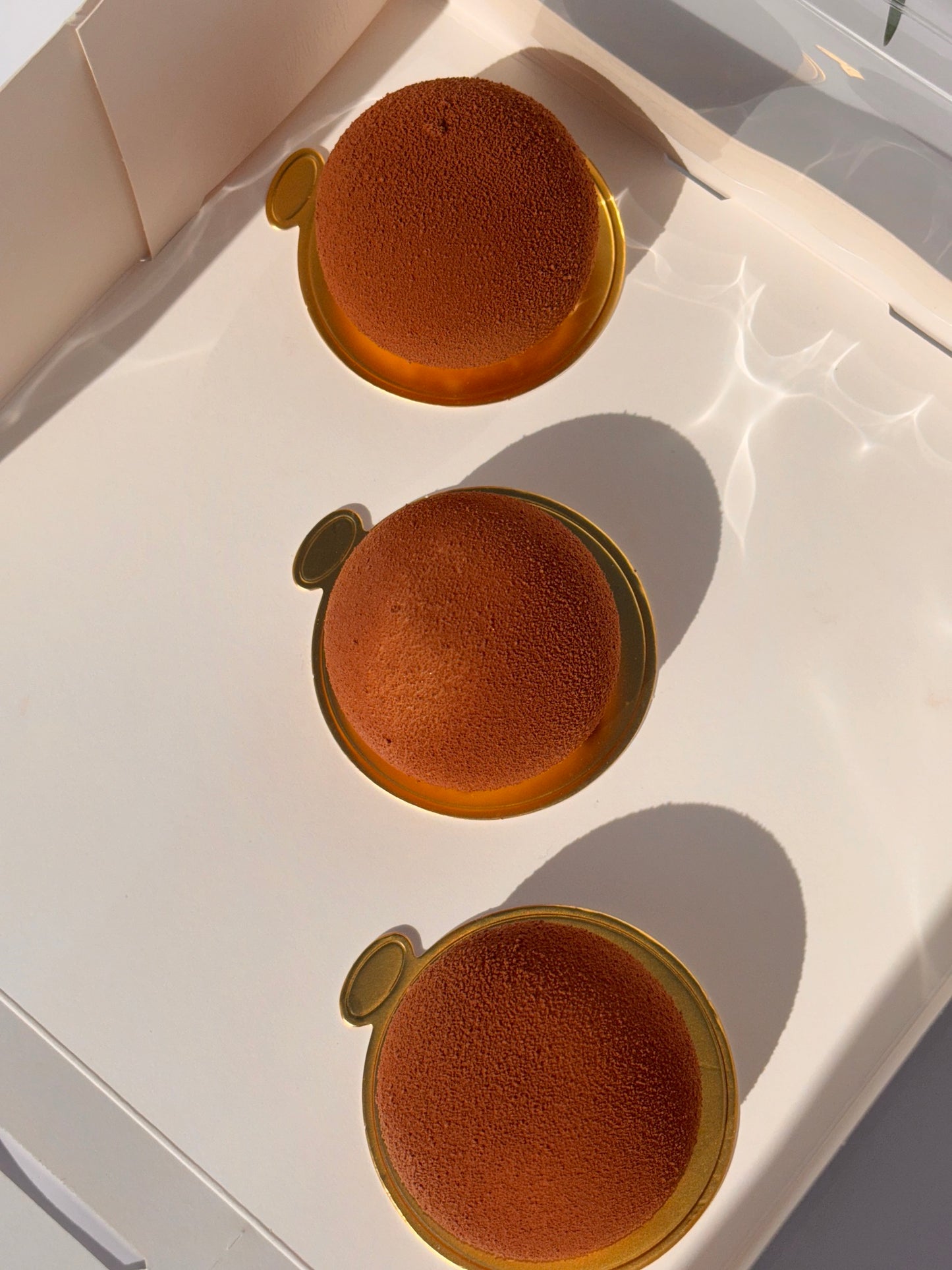 Tre bruna runda mini entremets med sammetsyta i en vit ask – Ganache Cakery Malmö.
Three brown round mini entremets with velvet texture in a white box – Ganache Cakery Malmö.
Trois mini entremets ronds bruns à texture veloutée dans une boîte blanche – Ganache Cakery Malmö.